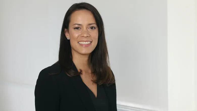 A dark-haired woman in a black suit stands before a white background 