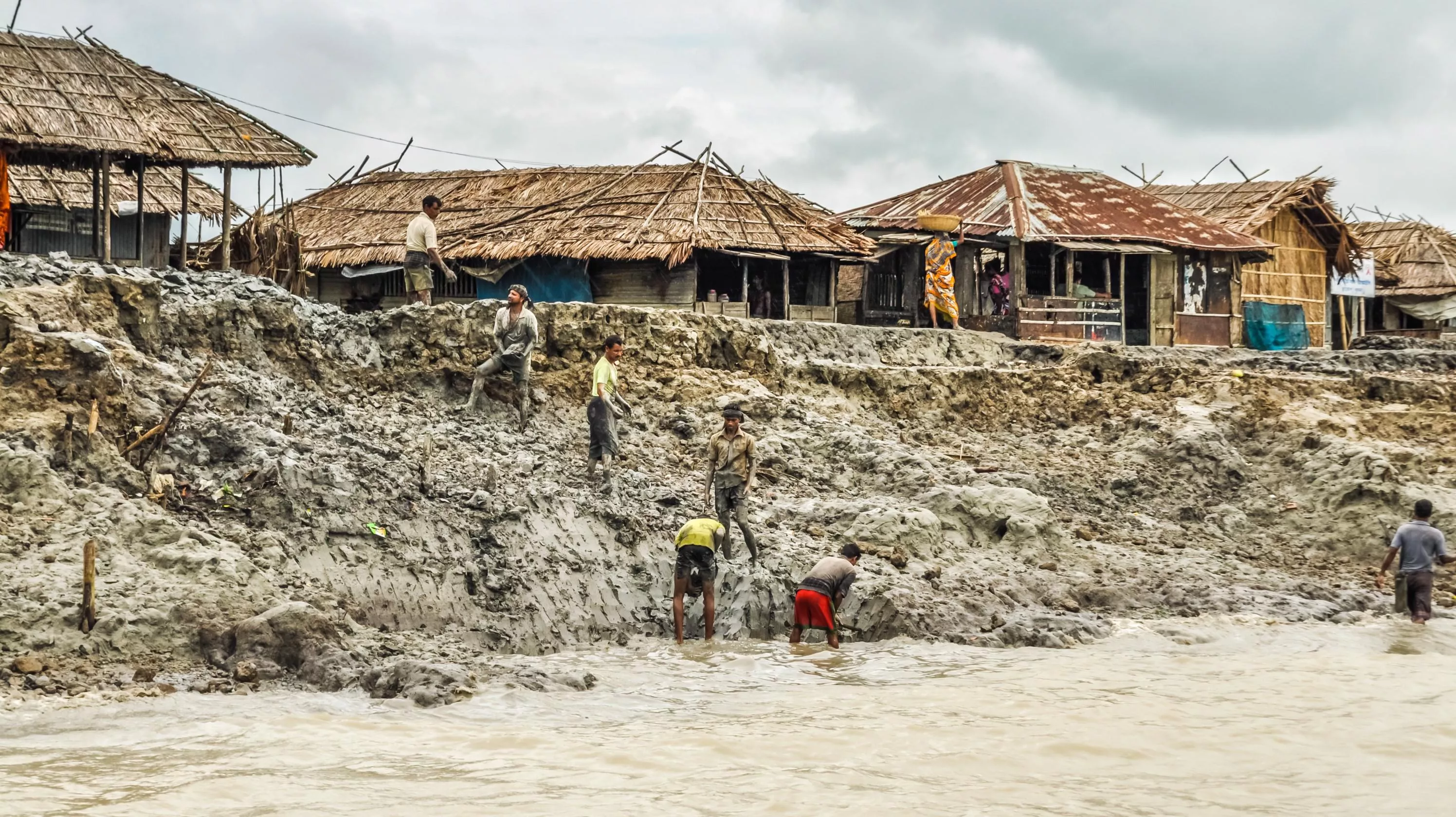 Men work on a muddy embankment with a river in the foreground and simple houses in the background.