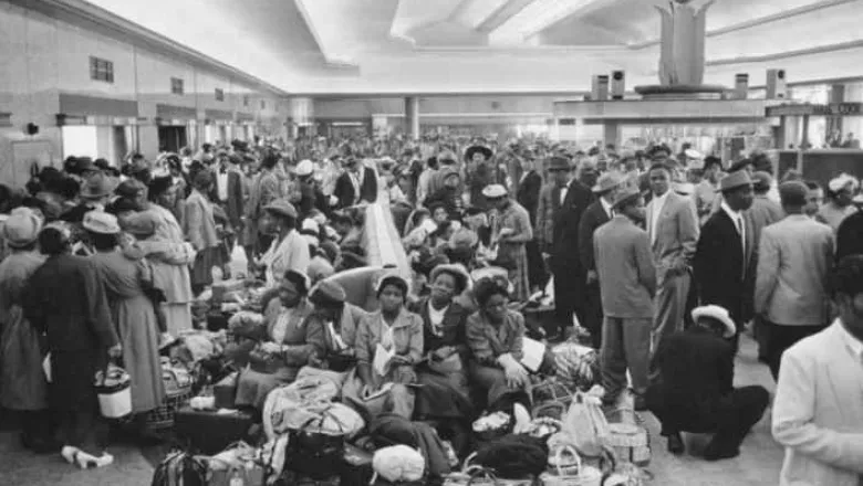 A black and white photo taken in the early 20th Century shows a group of men and women of afro-carribean descent waiting in a room. 