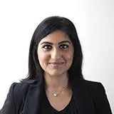 A woman with black hair and a black suit smiles against a white background headhot. 