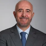 A man with a grey beard and dark eyebrows is wearing a gray suit with a blue tie and smiles against a gray background 