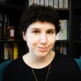 A woman with short dark hair is looking to the camera. There are books behind her. 