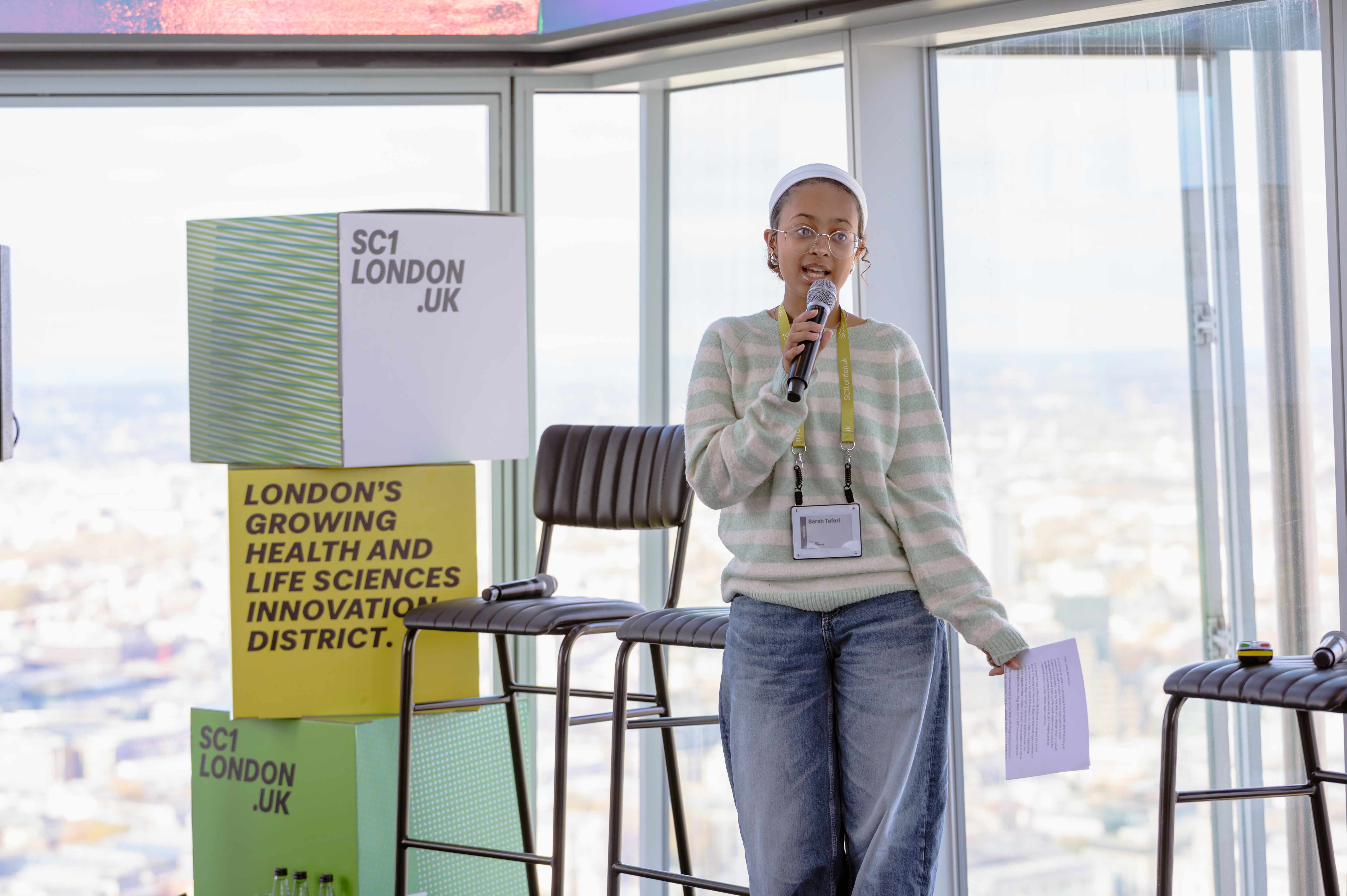 Sarah, wearing a green striped jumper and jeans, speaks into a microphone on a stage.