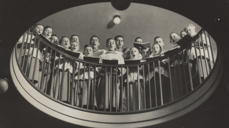 Choir in robes singing from a circular balcony at the Bear Pit in King’s, viewed from below.