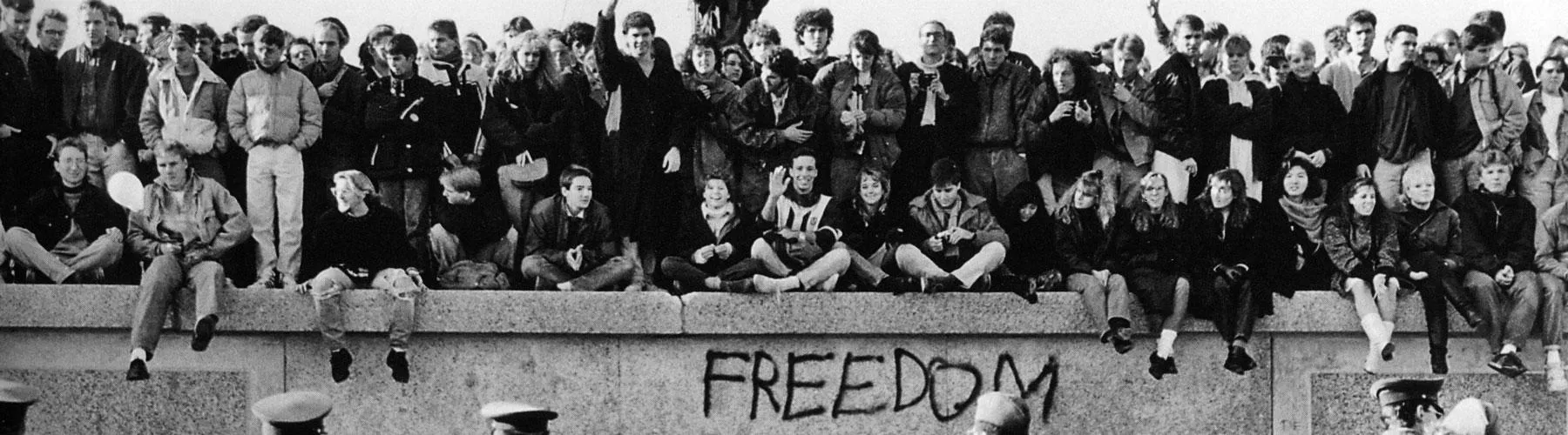 East German students sit atop the Berlin Wall at the Brandenburg Gate in front of border guards