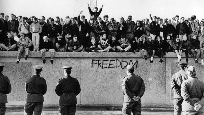 East German students sit atop the Berlin Wall at the Brandenburg Gate in front of border guards