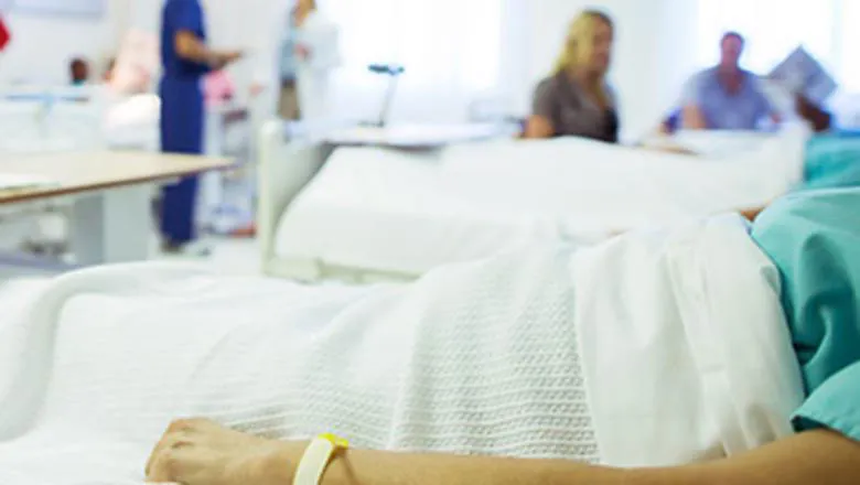 A patient lies in a hospital bed, their head is out of frame, healthcare staff in different uniforms work in the background