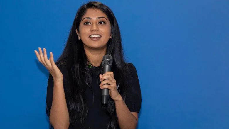 A woman in a black dress talks into a microphone, making a hand gesture of expression