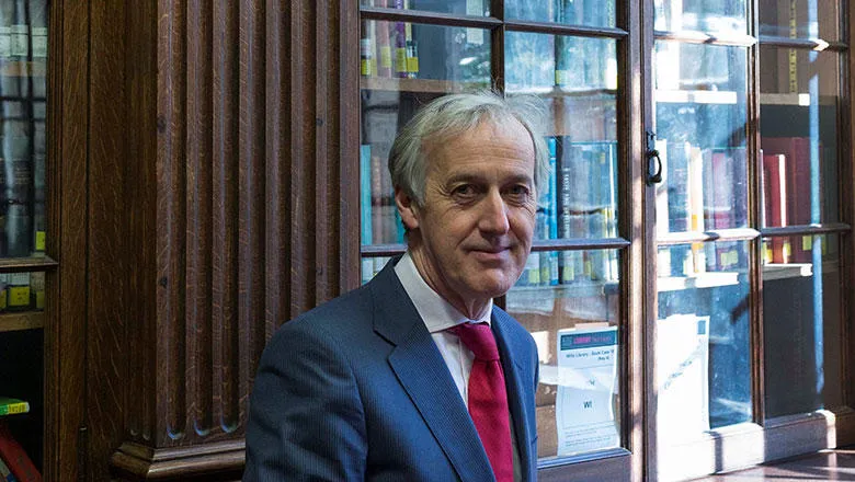 Professor Richard Trembath stood in front of display cabinet containing books.