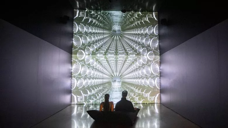 Two people sit on a beanbag in front of a geometric artwork in a dark room