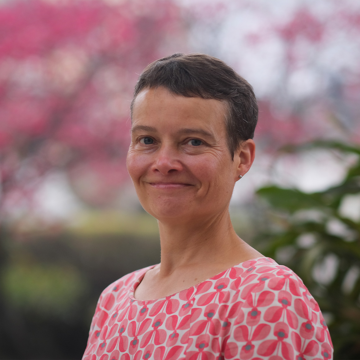 Melanie Barlow, a person with short dark hair, dressed in a pink shirt, standing in front of a cherry blossom tree.