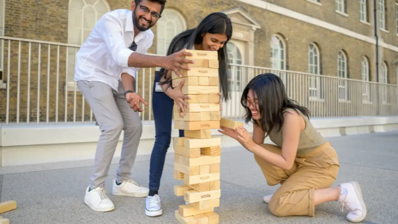 Three students play giant Jenga outside a historic building, laughing as the tower wobbles.