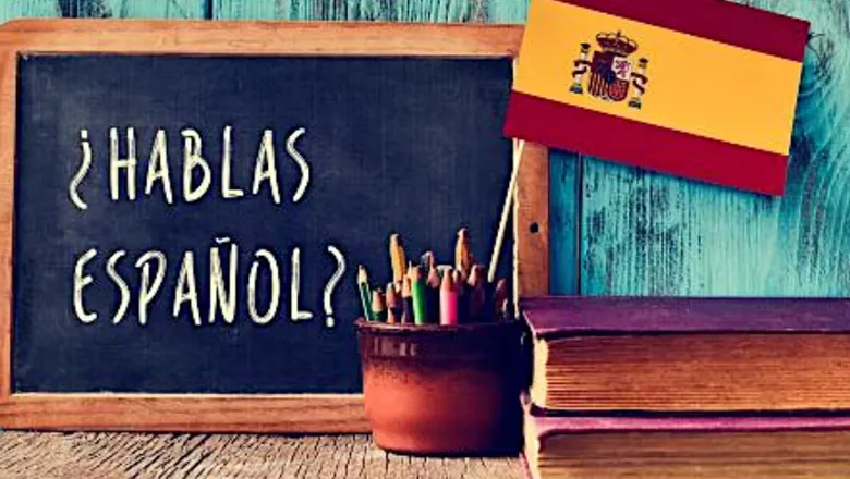 Picture of a Spanish flag sticking out of a pencil pot in front of a blackboard with Spanish text, and two books to the right.