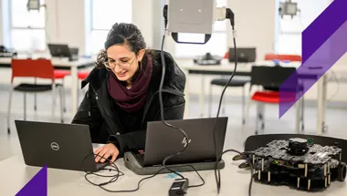 A young women works on two computers while a robotic device sits next to her.