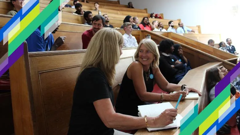 Two teachers talk to each other, sitting on wooden benches in a lecture theatre.