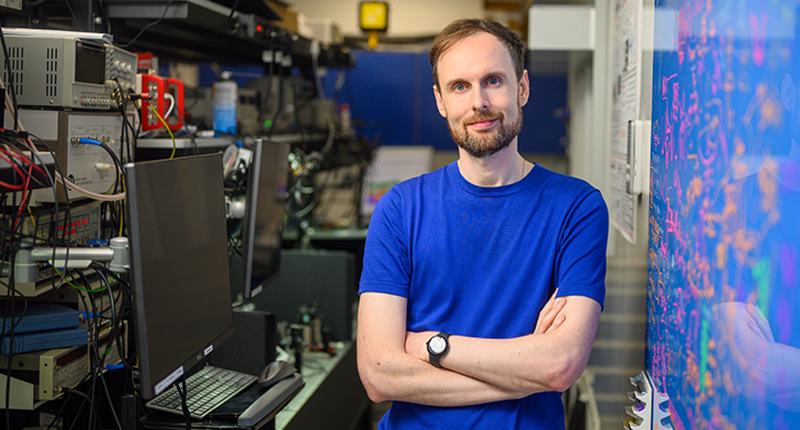 Professor James Millen stands in laboratory with lasers and equations on blueboard in background