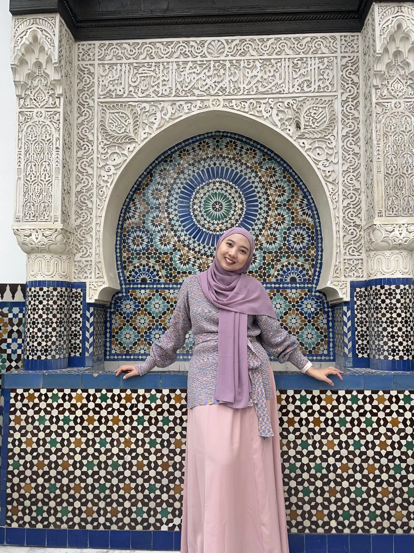 A woman wearing a pink hijab posing in front of a wall featuring an intricate and colorful tile pattern
