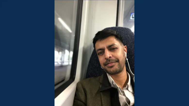 A selfie of Rasheed on a train with headphones in wearing a suit. The selfie is flanked by two midnight blue columns