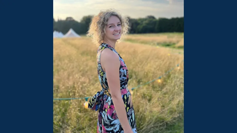 A woman with short curly blonde hair stands smiling in a field at sunset.