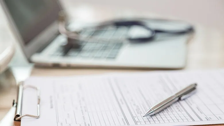 Stethoscope laid on laptop keyboard next to a paper form on a clipboard and a pen