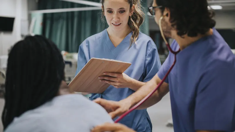 Patient having their chest heard by a doctor and a nurse is standing beside them writing notes.
