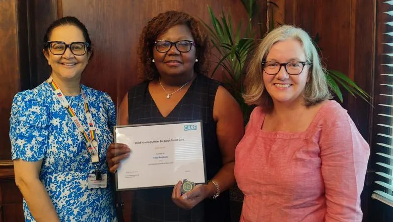 Three people pose for a photo in a wood panneled room, one in the middle holds up a certificate.