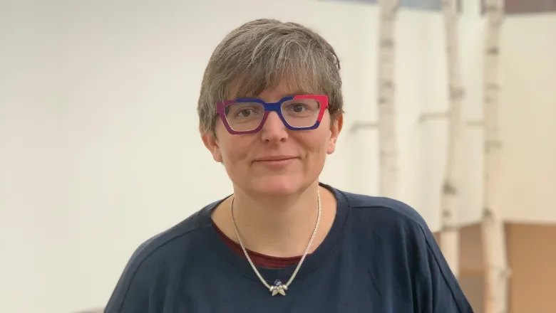 Person with cropped hair and multicoloured glasses smiles, wearing a long sleeved navy top.