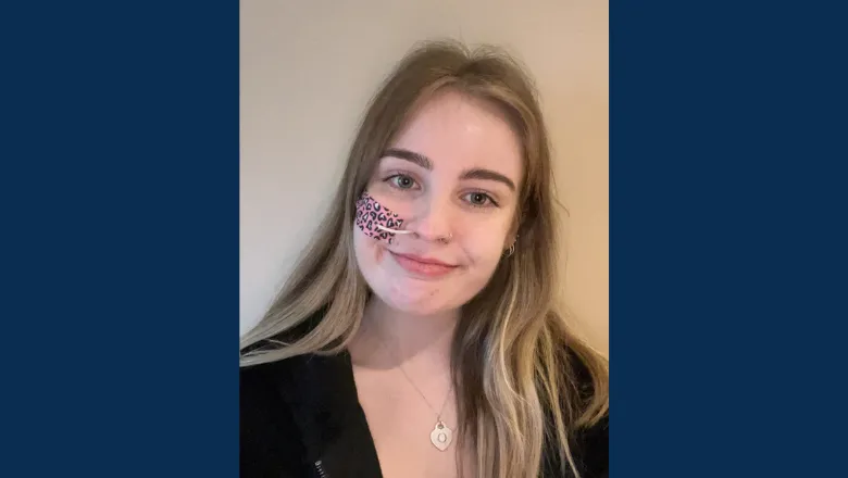A person with long blonde hair smiles at the camera. They have pink leopard print tape on their face keeping a breathing tube in place.