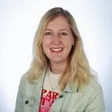 Blonde woman facing camera and smiling wearing light denim jacket and white t shirt, in front of a white blurred background.