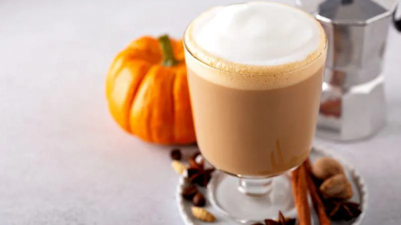 Frothy pumpkin spice latte in a clear glass, surrounded by a pumpkin, cinnamon sticks, and spices on a light surface. Cozy autumn vibe.