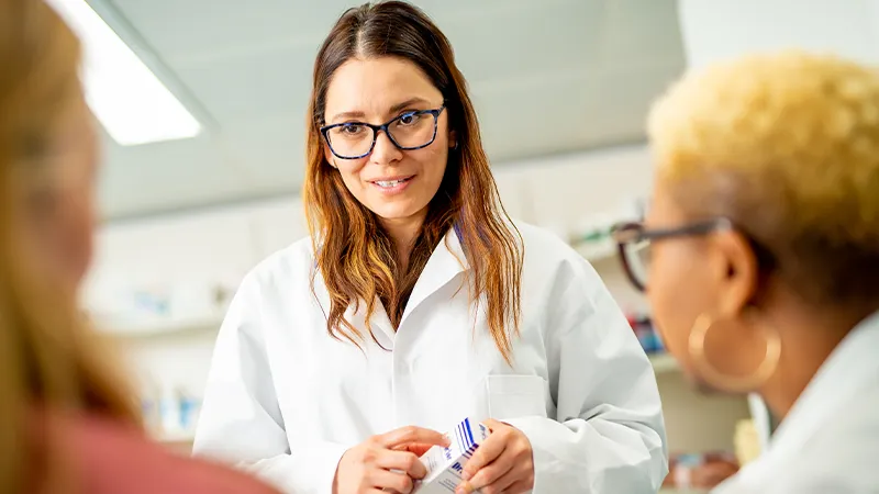 A person wearing glasses and a white lab coat holding a small box, in conversation with two other people