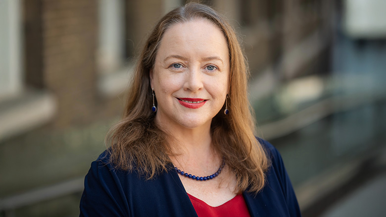 Bryony is a woman with red hair. She is wearing a red top and a blue blazer.