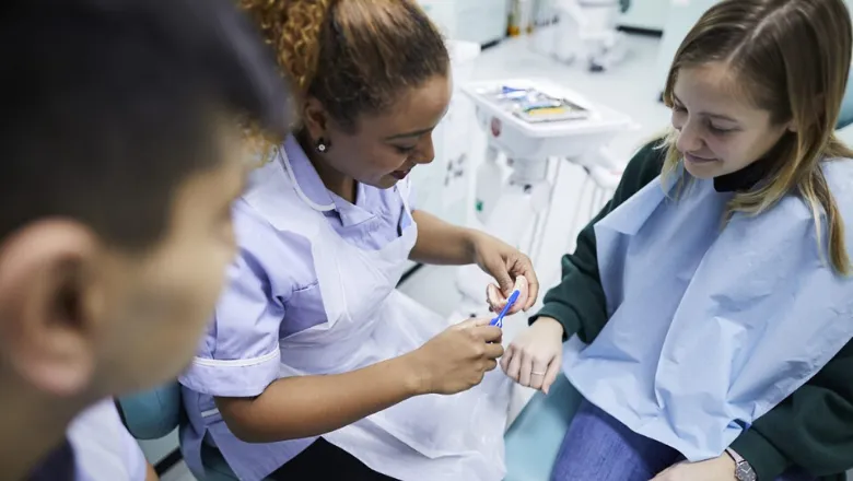 A person dressed in medical uniform shows a patient wearing a bib a model of a mouth and toothbrush