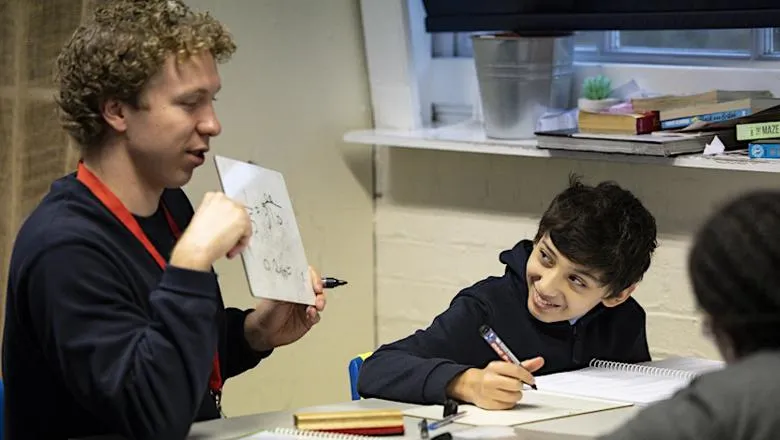 A boy on left holding a white paper tutoring a boy on the right 