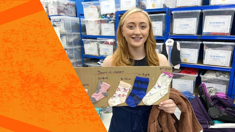 A young woman with blonde hair is holding some cardboard that shows the various sizes of baby socks - it has text thats says: Sock-o-meter. In the background there is a number of different boxes of donations.