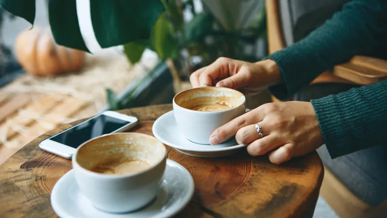 Two cups of coffee on a table with one individuals hands holding a cup