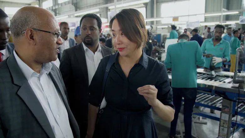 A man and woman standing together within a large factory building, both looking at each other with neutral expressions.