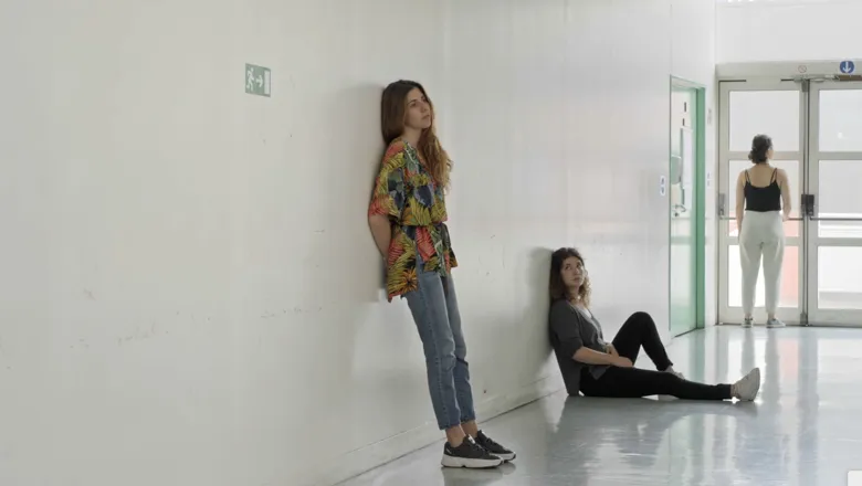 Image of 2 women in an empty university hall, one leaning against a wall, one person sitting down