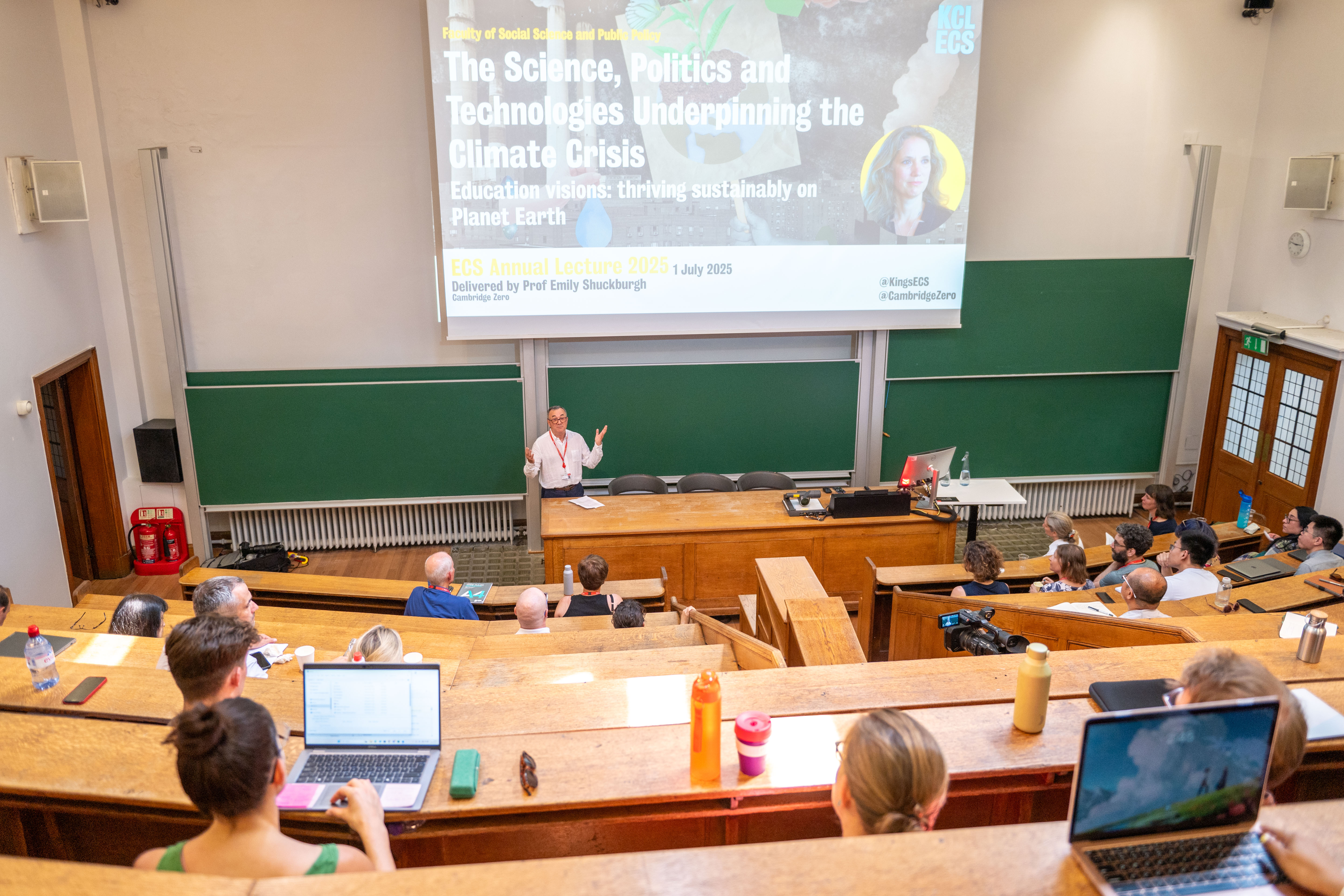 far away shot of the lecture theatre, people with laptops and a male at the front under a projector screen
