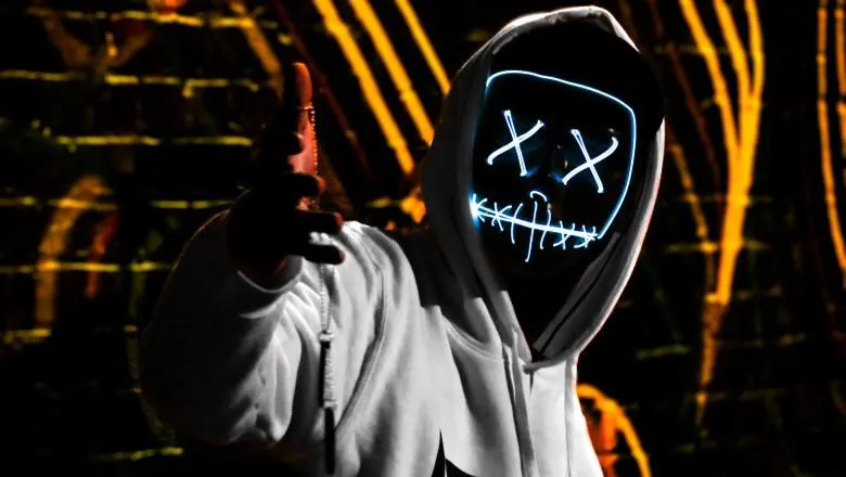 A man is lifting his right hand a bit like a gun gesture, he wears a grey hoodie and has a mask on with Xs in place of eyes.