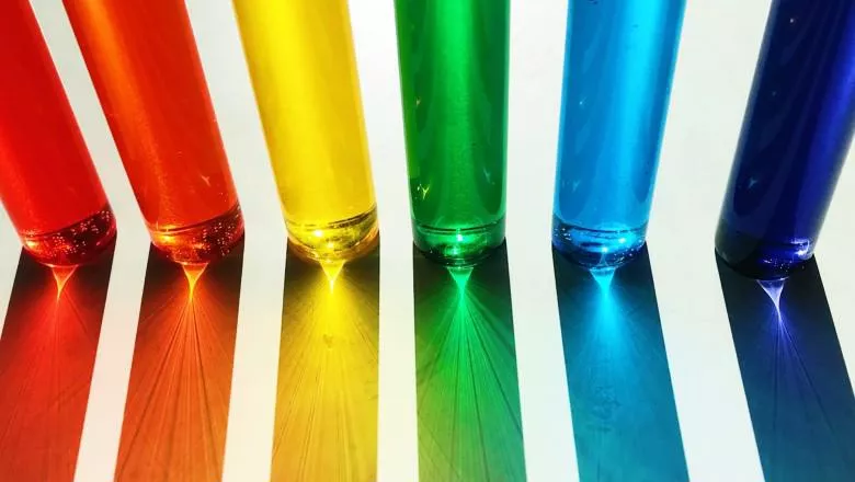 Photo of 6 narrow glasses next to each other, representing the rainbow in the colour of their respective liquid - from left to right: red, orange, yellow, green, sky blue, dark blue.
