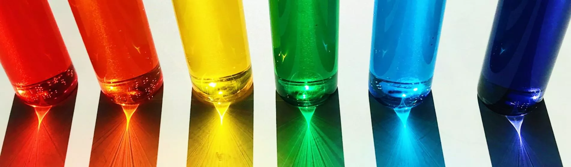 Photo of 6 narrow glasses next to each other, representing the rainbow in the colour of their respective liquid - from left to right: red, orange, yellow, green, sky blue, dark blue.