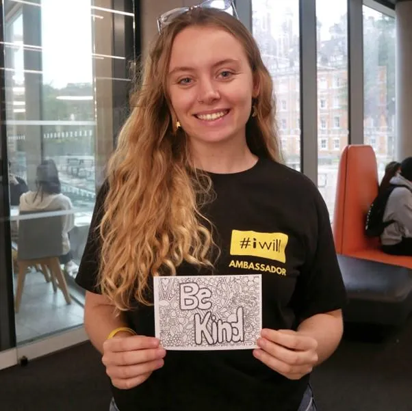 woman with long gingery blonde hair holds a sign that says 'be kind'