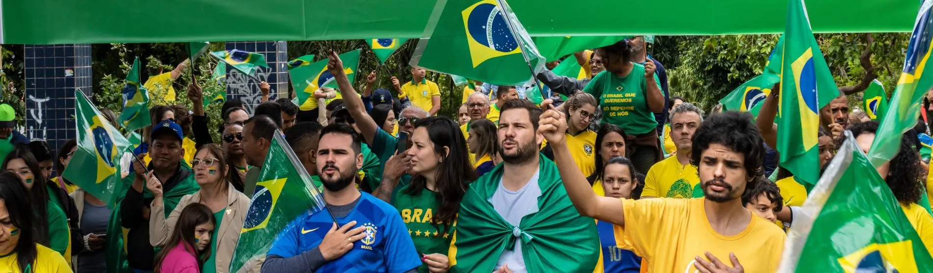 Protesters waving the Brazilian flag, with three men in the foreground holding their hand over their heart and singing.