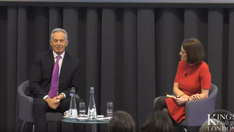 Former Prime Minister Tony Blair sits in a chair with a purple tie, as Rachel Sylvester in a red dress asks him a question.