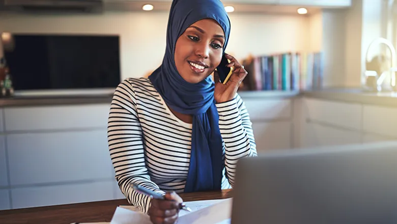 Woman in a headscarf with a phone to hear ear and a pen in hand, in a home office environment