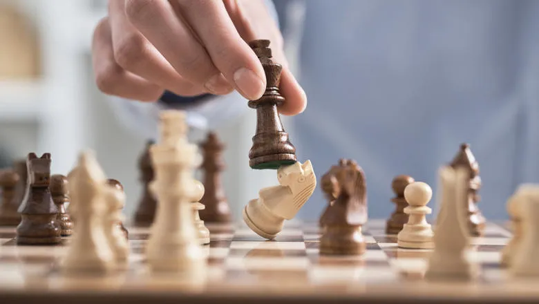 Hand holding a black king chess piece over a chess board, knocking over a white rook chess piece