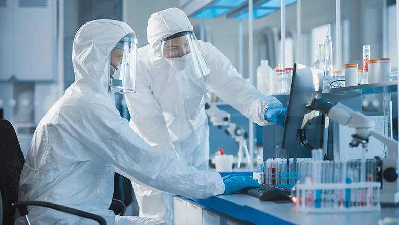 Two researchers dressed in protective equipment look at a computer in lab. 