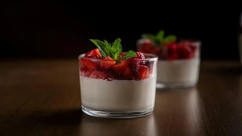 Two glass cups of creamy panna cotta topped with vibrant red strawberries and fresh mint leaves sit on a wooden table, set against a dark background.