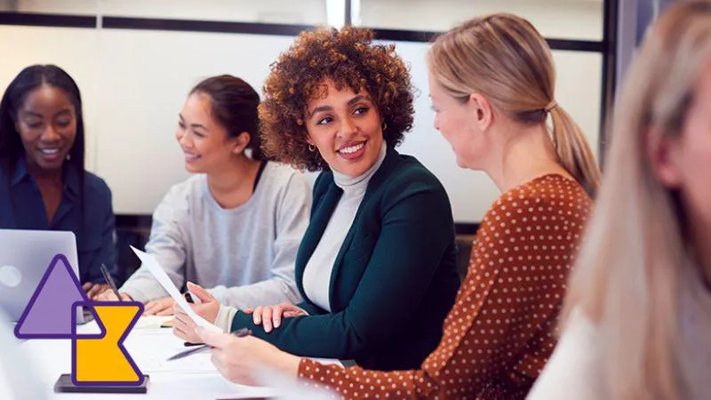 Image: Two women look at each as if discussing something, they both are other holding papers and sit in a larger group with laptops around them.